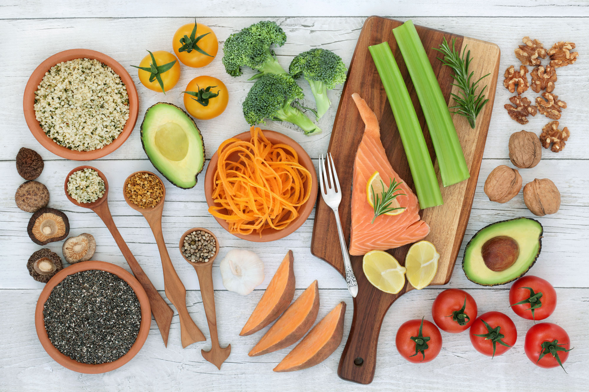 Raw salmon fillet on a wooden cutting board garnished with lemon and rosemary, surrounded by halved avocados, yellow and red tomatoes, broccoli, celery, spiralized carrots, sweet potato wedges, walnuts, garlic, mushrooms and bowls of seeds and grains on a white wooden surface.