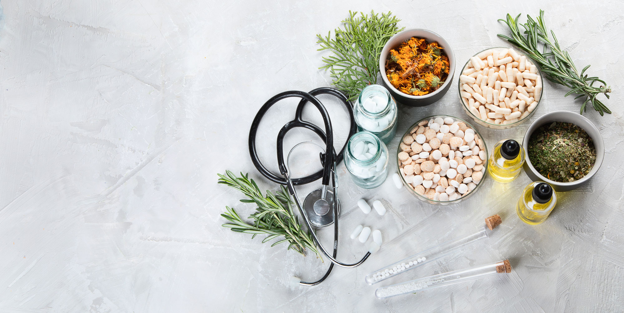 Black stethoscope placed among glass jars of tablets, bowls of capsules and dried herbs, small bottles of oil, rosemary sprigs and two corked test tubes on a pale textured surface, suggesting a mix of conventional and herbal remedies.