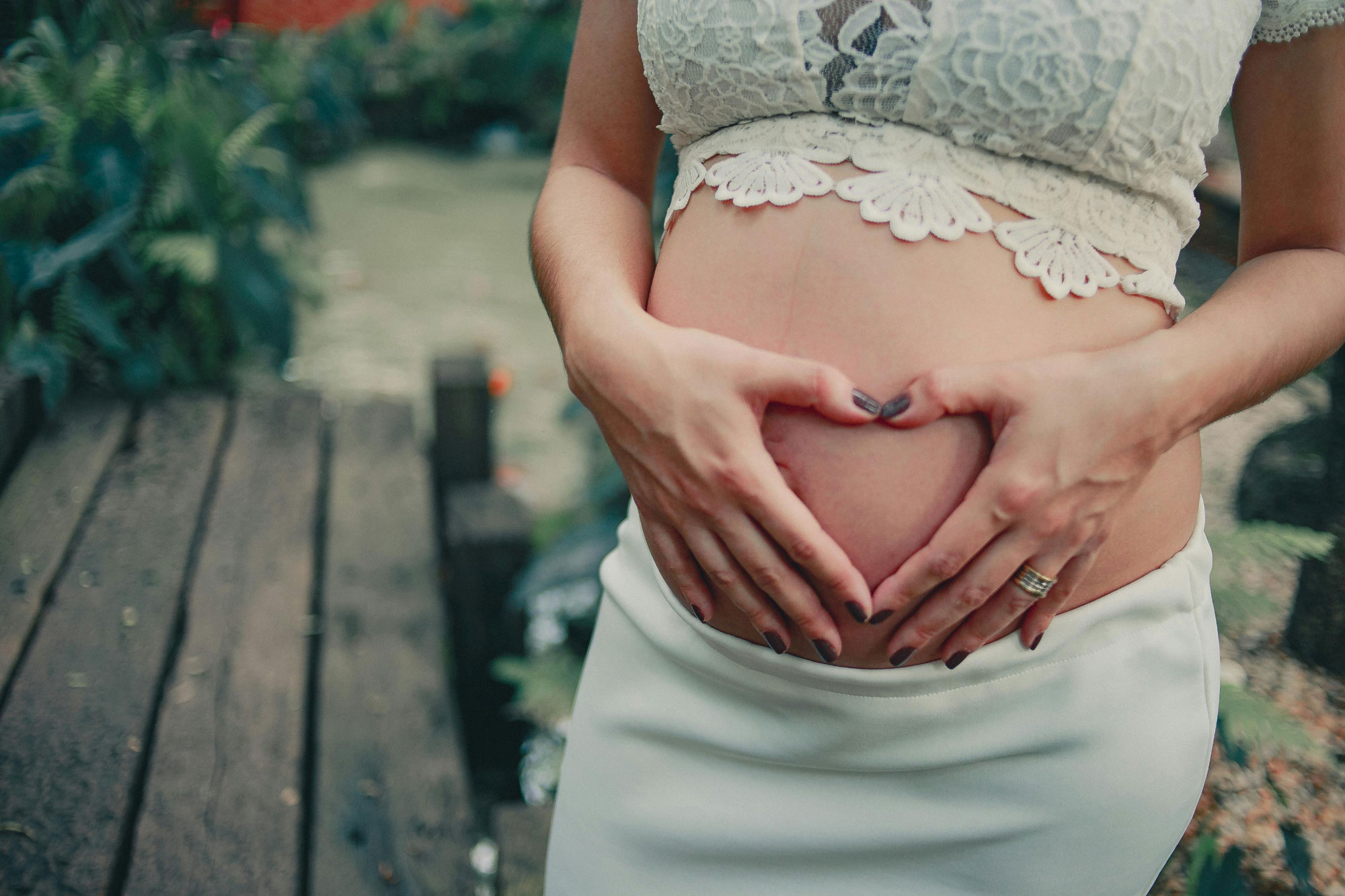 Pregnant woman's bare belly framed by hands forming a heart shape, wearing a white lace crop top and skirt with a wedding ring visible, standing outdoors on a wooden deck with blurred greenery behind her.
