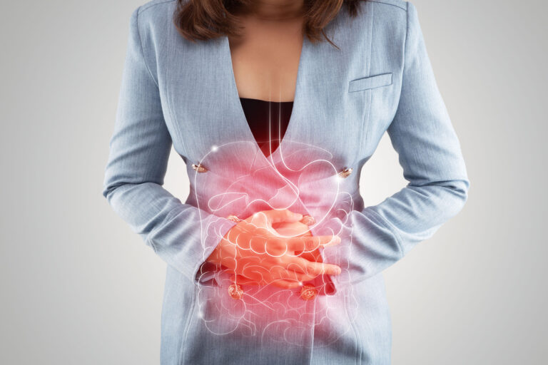 Woman in a light blue blazer clutching her lower abdomen with a translucent glowing red illustration of the stomach and intestines overlaid to indicate abdominal pain.