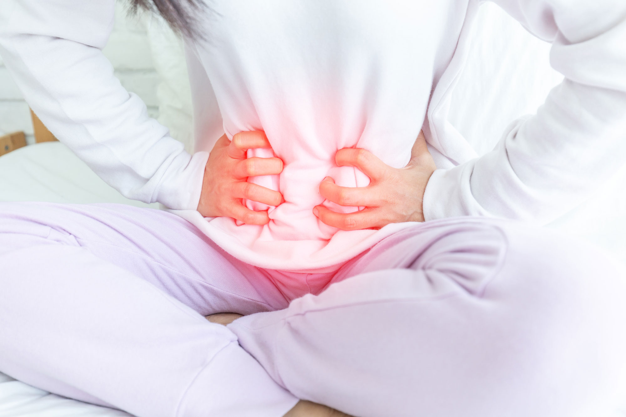 Person seated cross-legged with both hands pressing their lower abdomen through a white shirt, a red glow highlighting abdominal pain.