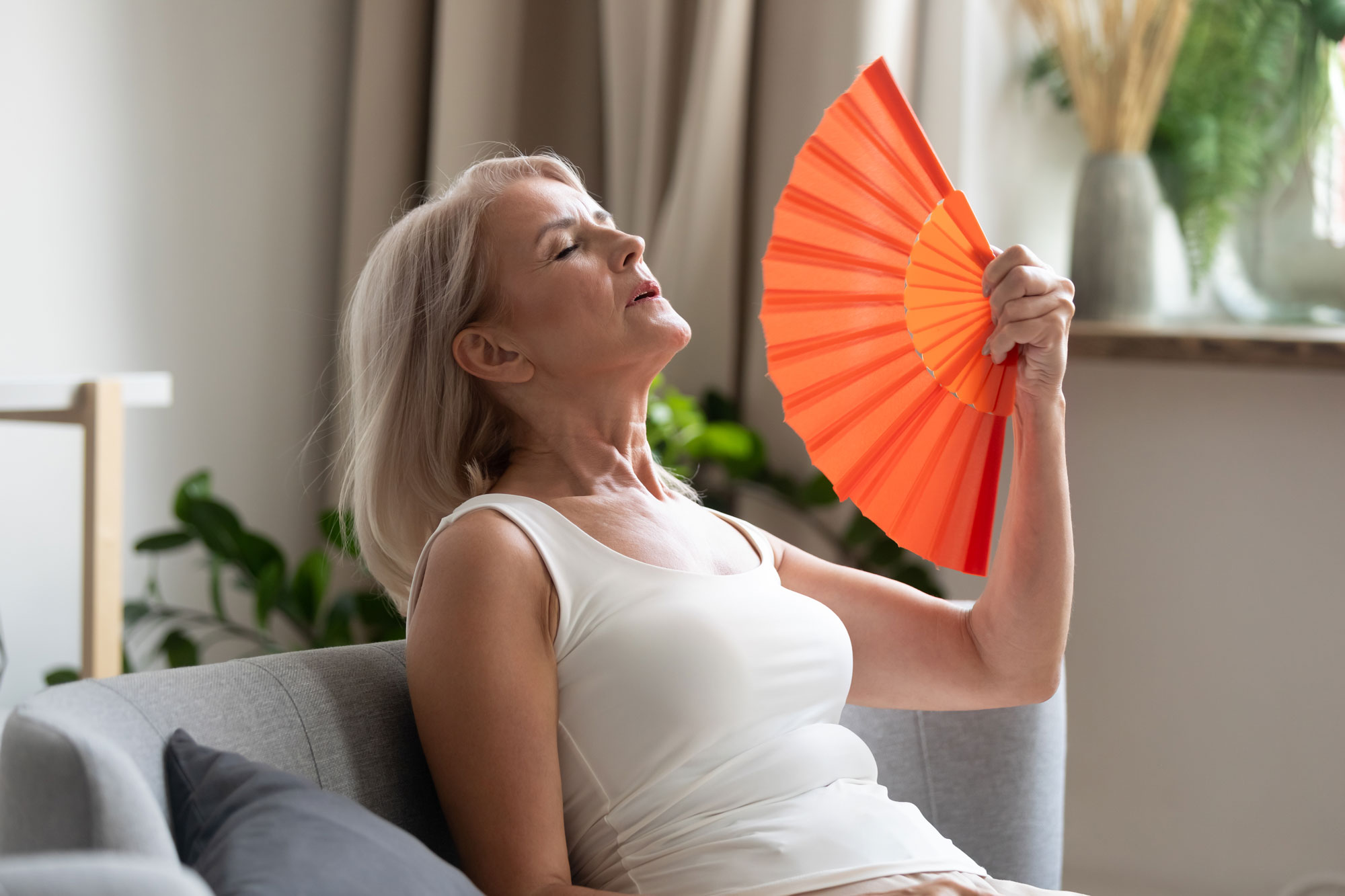 Older woman reclining on a gray sofa in a bright living room, wearing a white sleeveless top and fanning herself with a large orange folding fan while her eyes are closed.