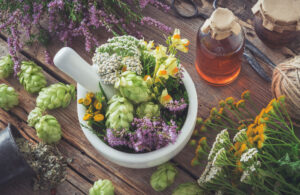 White ceramic mortar and pestle overflowing with green hop cones, white yarrow and yellow and purple wildflowers, set on a weathered wooden table with scattered hop cones, an amber jar, twine and bundles of herbs.