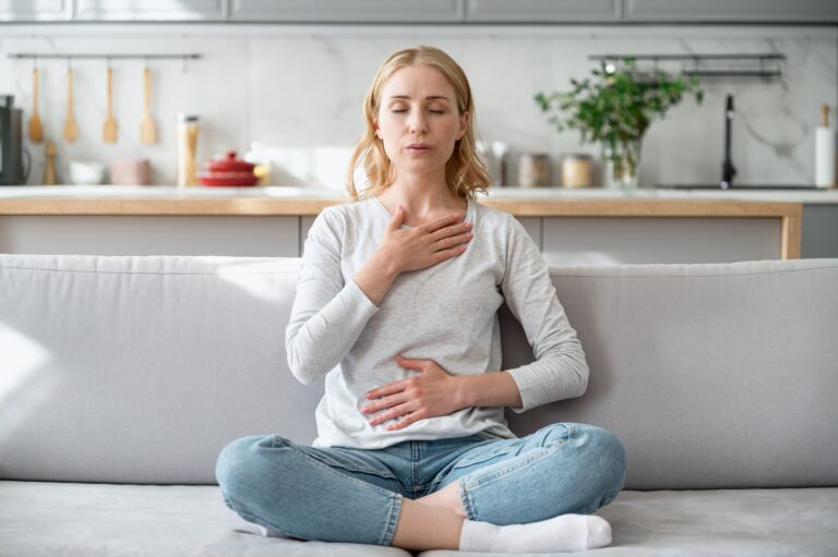 A blonde woman sits cross-legged on a gray couch in a bright modern kitchen, eyes closed with one hand on her chest and the other on her abdomen while practicing deep, calming breaths.