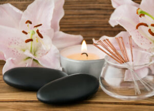 Two smooth black massage stones, a lit tealight candle, a glass jar holding copper-handled acupuncture needles, and pale pink lily blossoms arranged on a wooden surface.