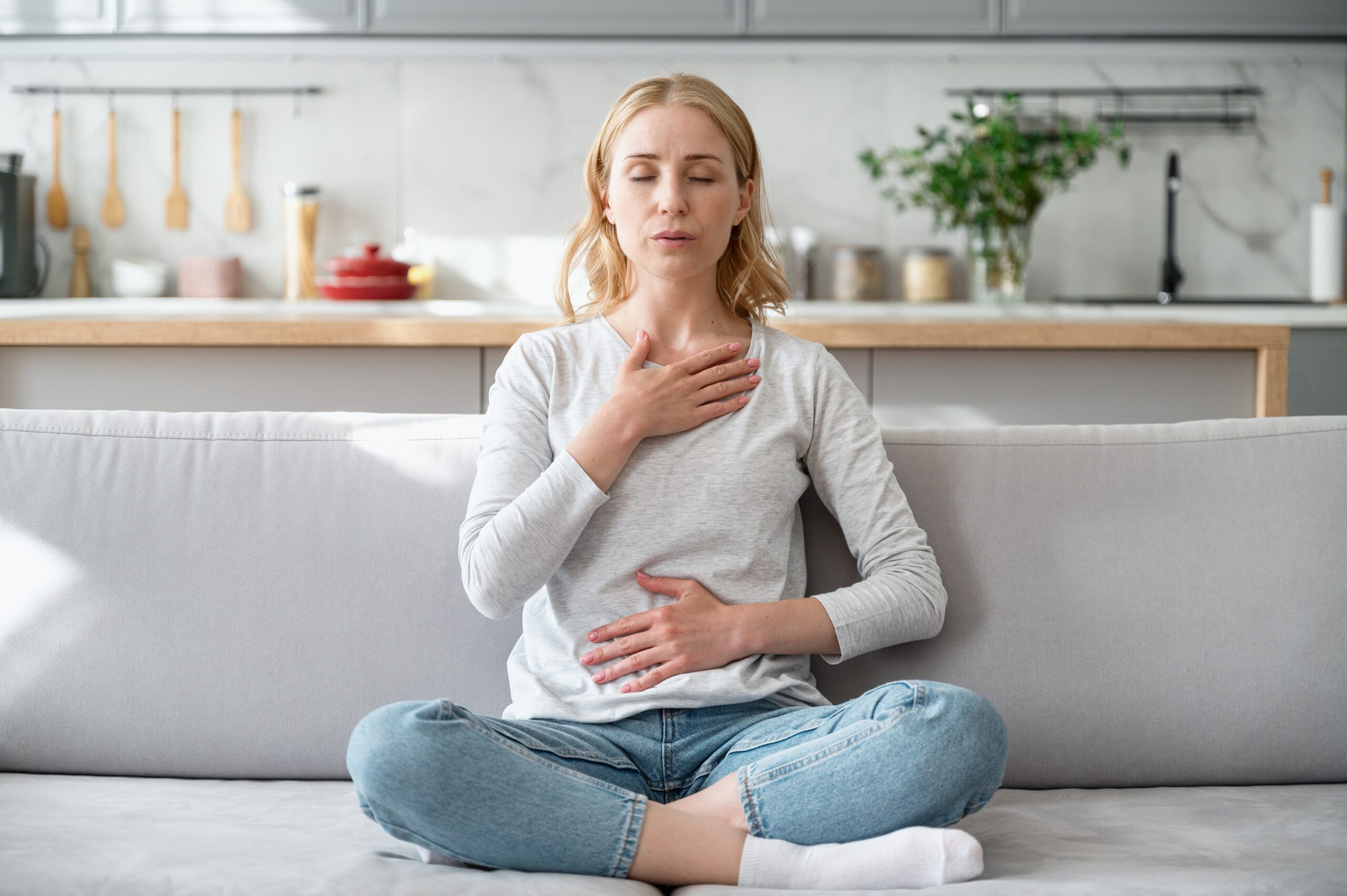 A blonde woman sits cross-legged on a gray couch in a bright modern kitchen, eyes closed with one hand on her chest and the other on her abdomen while practicing deep, calming breaths.