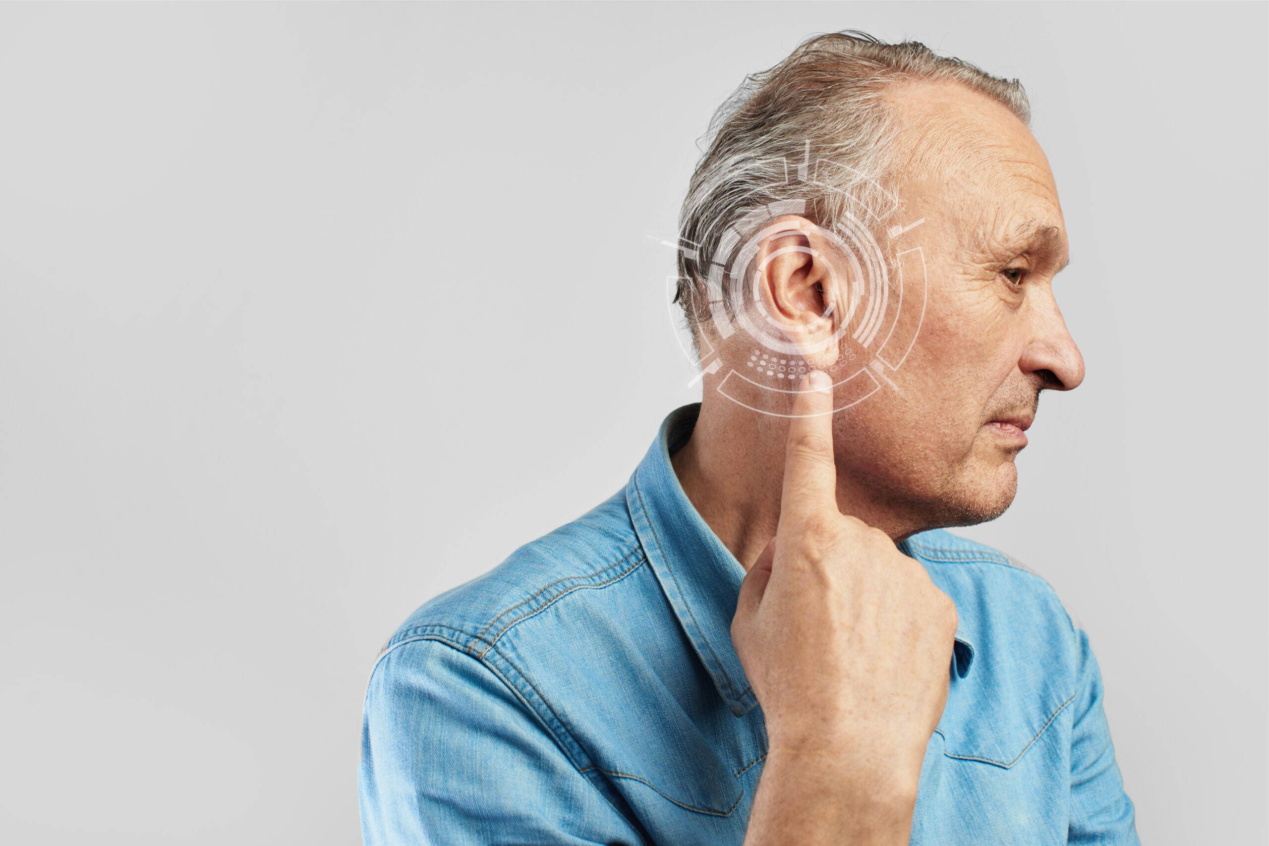 Older man in a blue denim shirt shown in profile, pointing to his ear with a translucent circular digital interface overlay around the ear suggesting hearing technology, on a plain light-gray background.