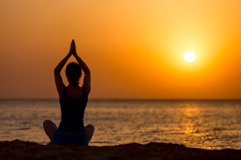 Silhouette of a person seated cross-legged on a beach with hands pressed together above their head in a yoga pose as the orange sun sets and reflects on the calm sea.