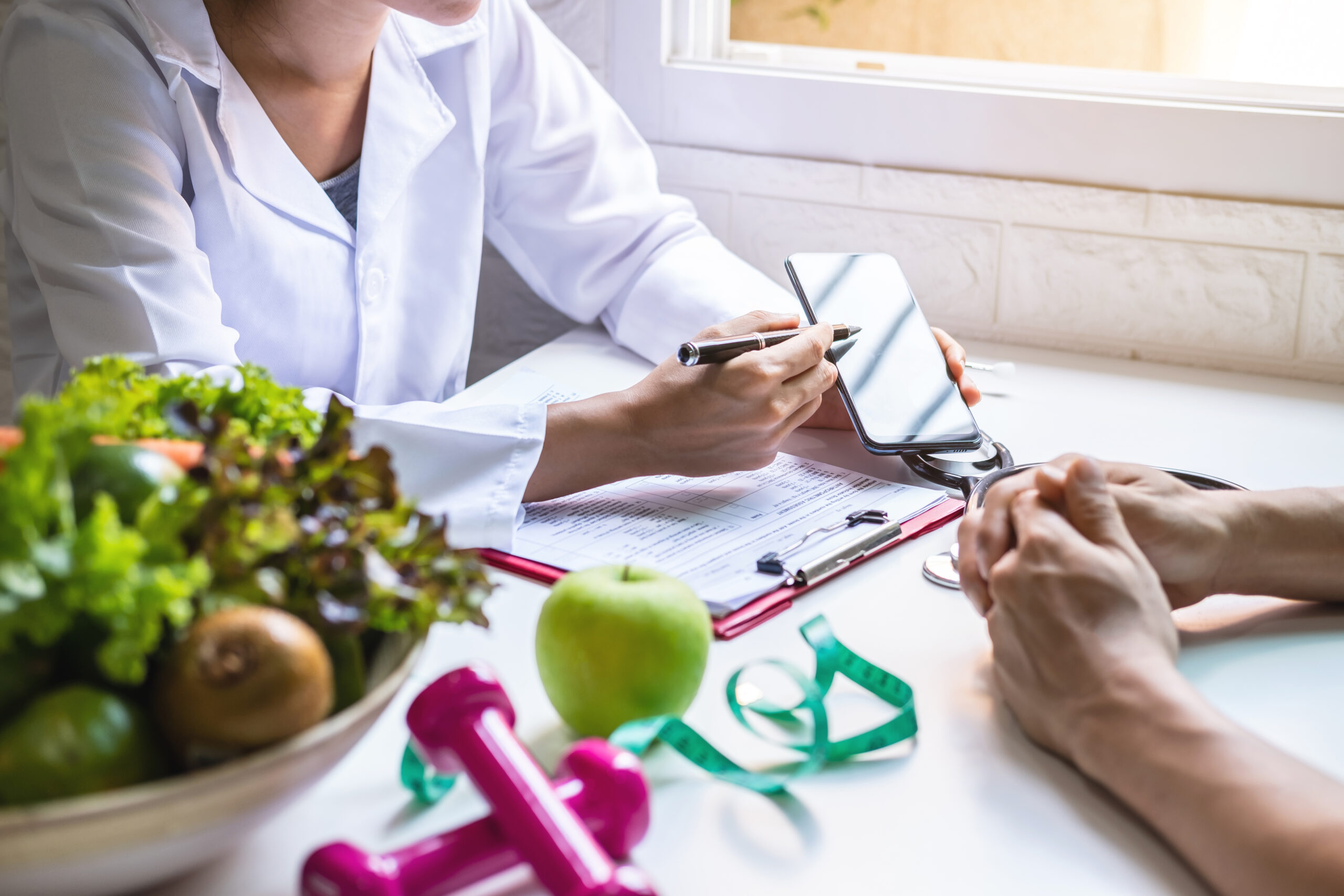 A healthcare professional in a white coat points to a smartphone during a consultation across a desk with a clipboard and a patient's clasped hands, while a bowl of salad, a green apple, a measuring tape and pink dumbbells in the foreground suggest nutrition and fitness advice.